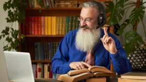 Distance Learning Concept. Teacher professor tutor in headphones teaches discipline online. Mature bearded man answers teacher’s question through laptop.