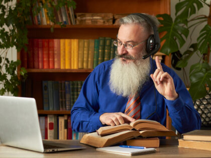 Distance Learning Concept. Teacher professor tutor in headphones teaches discipline online. Mature bearded man answers teacher’s question through laptop.