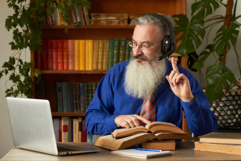 Distance Learning Concept. Teacher professor tutor in headphones teaches discipline online. Mature bearded man answers teacher's question through laptop.